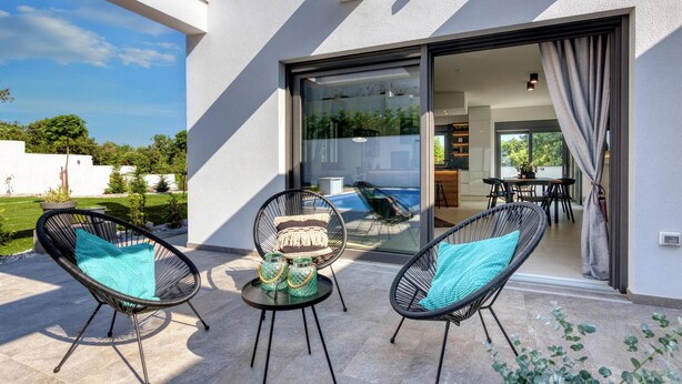 A modern outdoor patio with two wicker chairs, a small round table, and a glass door leading to a kitchen and dining area.