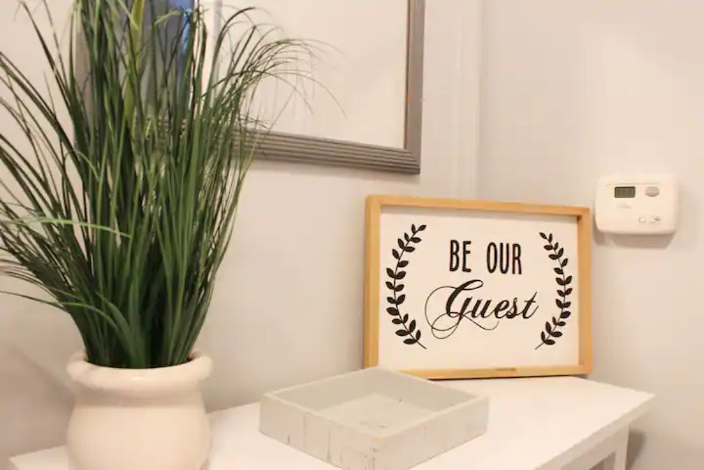 A framed sign with the text "BE OUR GUEST" surrounded by a decorative wreath, placed on a white surface next to a potted plant.
