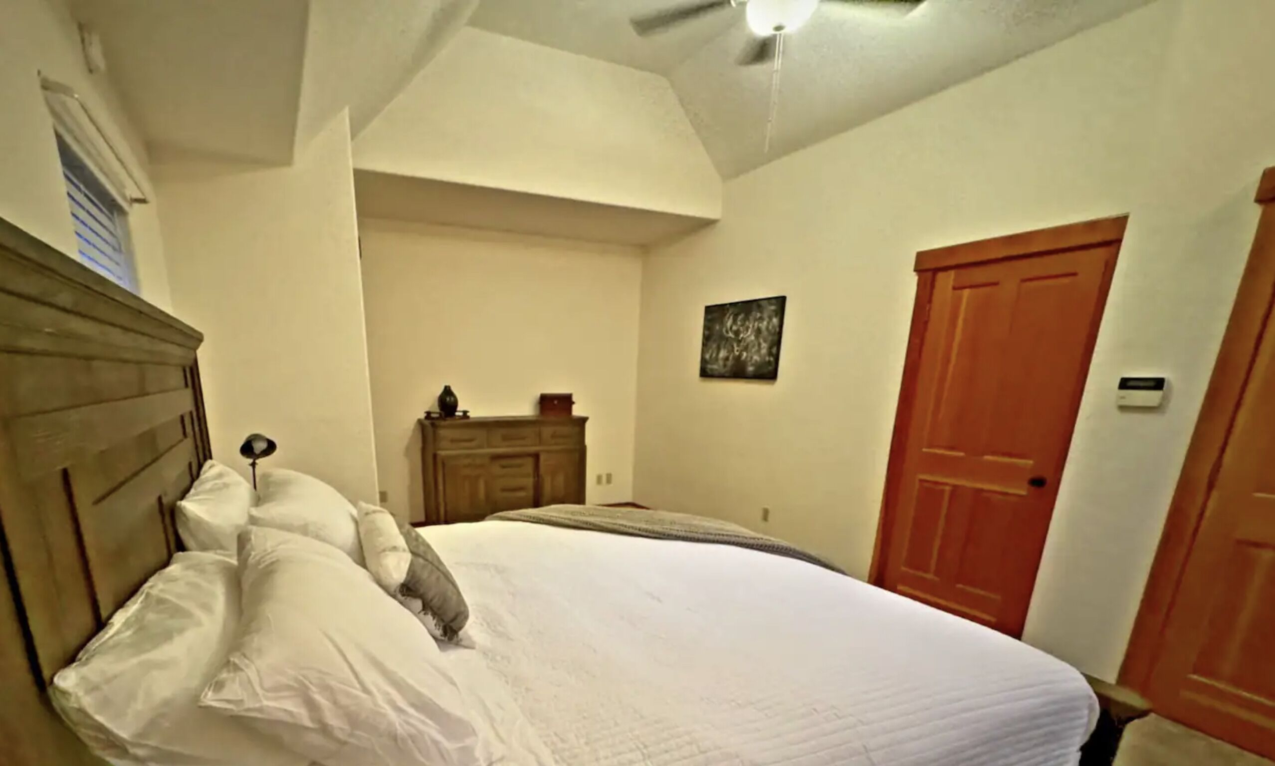 A bedroom with a wooden headboard, a bed with white bedding, a dresser, a door, and a ceiling fan.