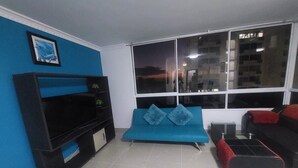 Ein Wohnzimmer mit blauem Sofa, schwarzem Sofa, Fernseher auf einem Schrank und einem großen Fenster mit Blick auf die Stadt.