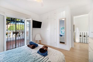 Un dormitorio con cama, televisor, una silla blanca y un balcón con vistas.