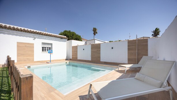 A swimming pool with lounge chairs, a wooden deck, and a white building with a tiled roof.