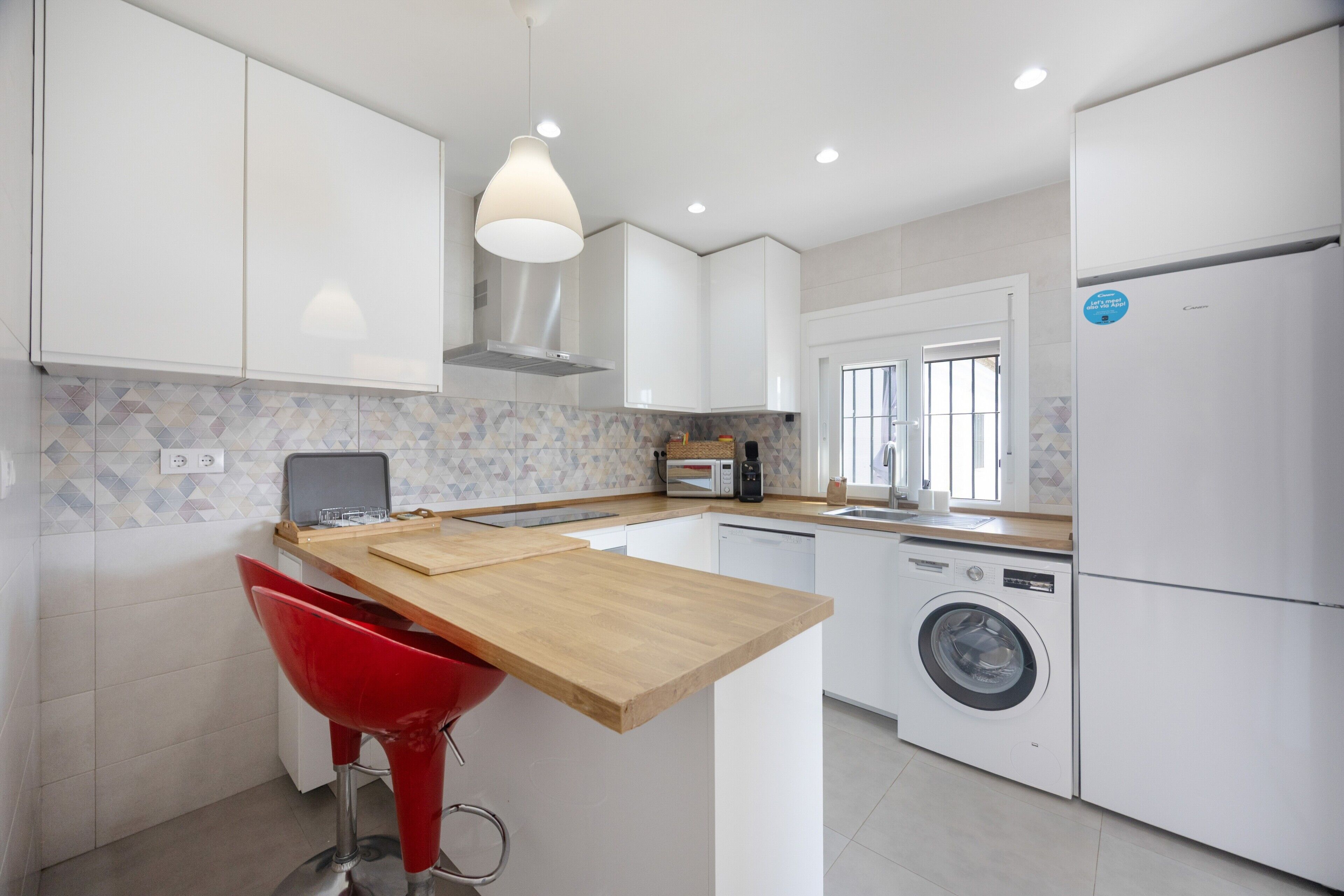 Una cocina moderna con armarios blancos, una isla de madera, una lavadora y una silla roja.