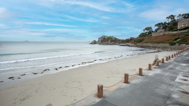 A beach with a concrete walkway, a stone wall, and a building on a hill. - Villa 'Bellevue Rue De La Petite Corniche' with Sea View, Private Terrace and Wi-Fi (perros guirec)