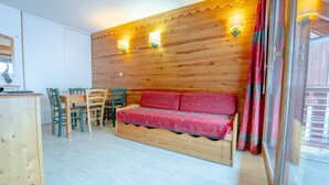 Eine Holzwand mit integrierter Beleuchtung, ein Holzsofa mit roter Auflage, ein Esstisch mit Stühlen und ein Fenster mit Vorhängen.