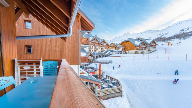 Ein Holzbau mit Balkon, der einen verschneiten Hang mit Skipisten und Gebäuden im Hintergrund überblickt.