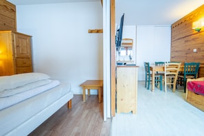 Quarto com cama, uma mesinha, armário de madeira, televisão e espaço de refeições com cadeiras.