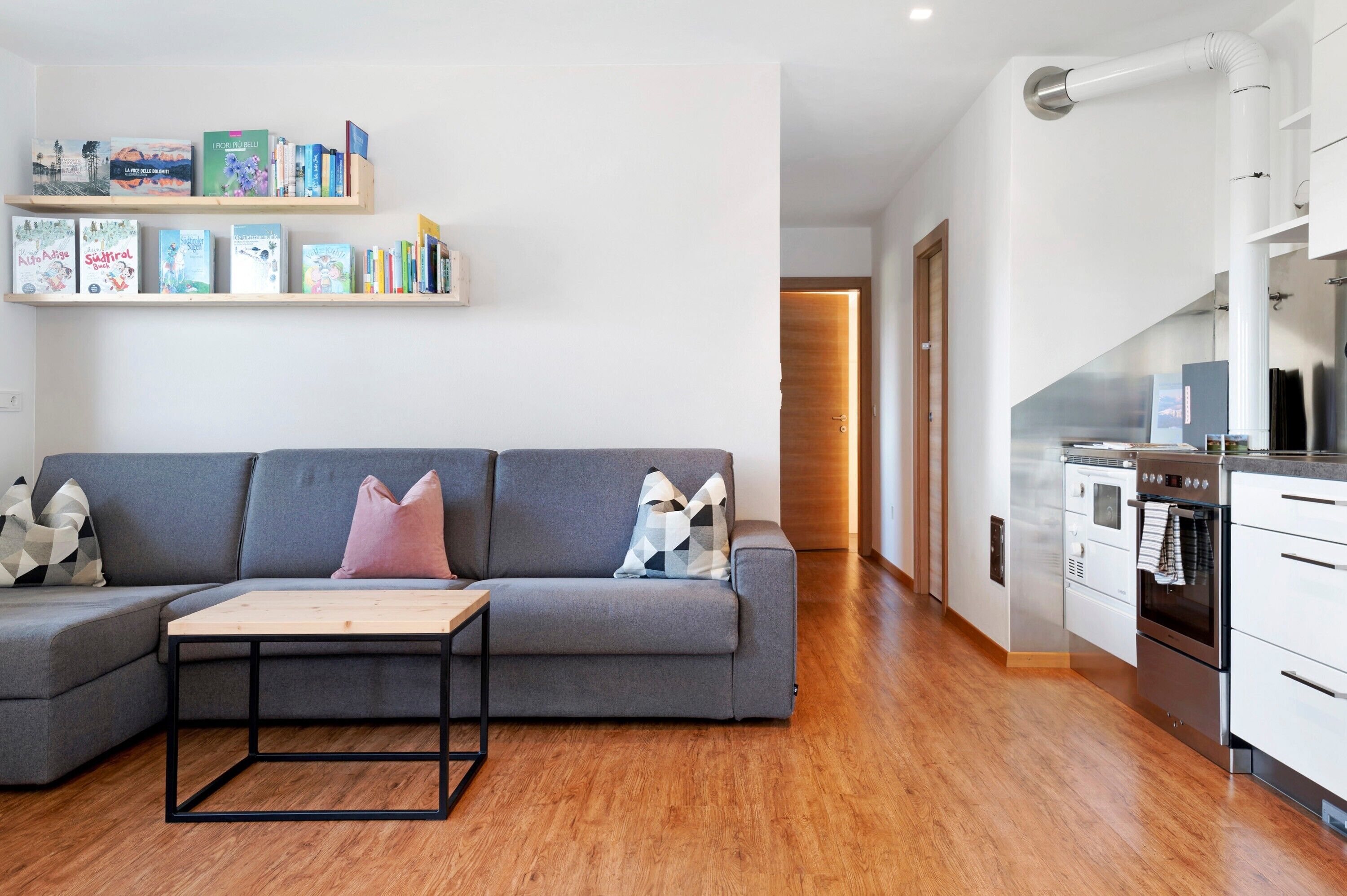 Ein modernes Wohnzimmer mit einem grauen Sofa, einem Holztisch und einer Küche inklusive Herd und Backofen.