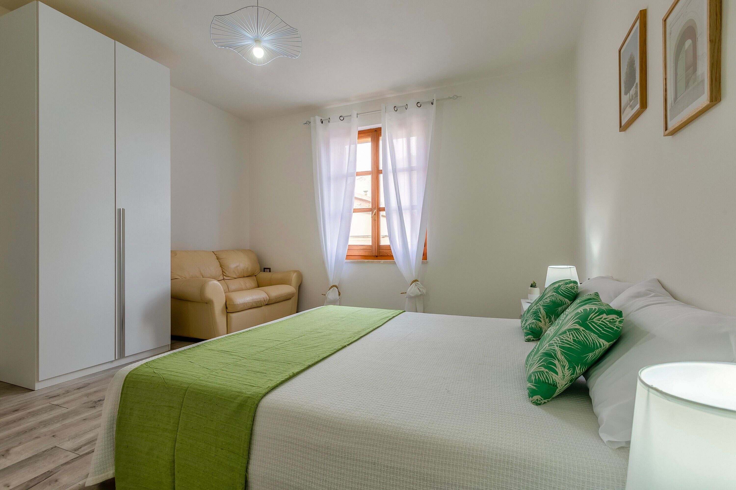 Une chambre à coucher avec un lit, un plaid vert, une chaise, une table de chevet et une fenêtre avec des rideaux.