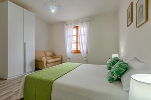 Une chambre à coucher avec un lit, un plaid vert, une chaise, une table de chevet et une fenêtre avec des rideaux.