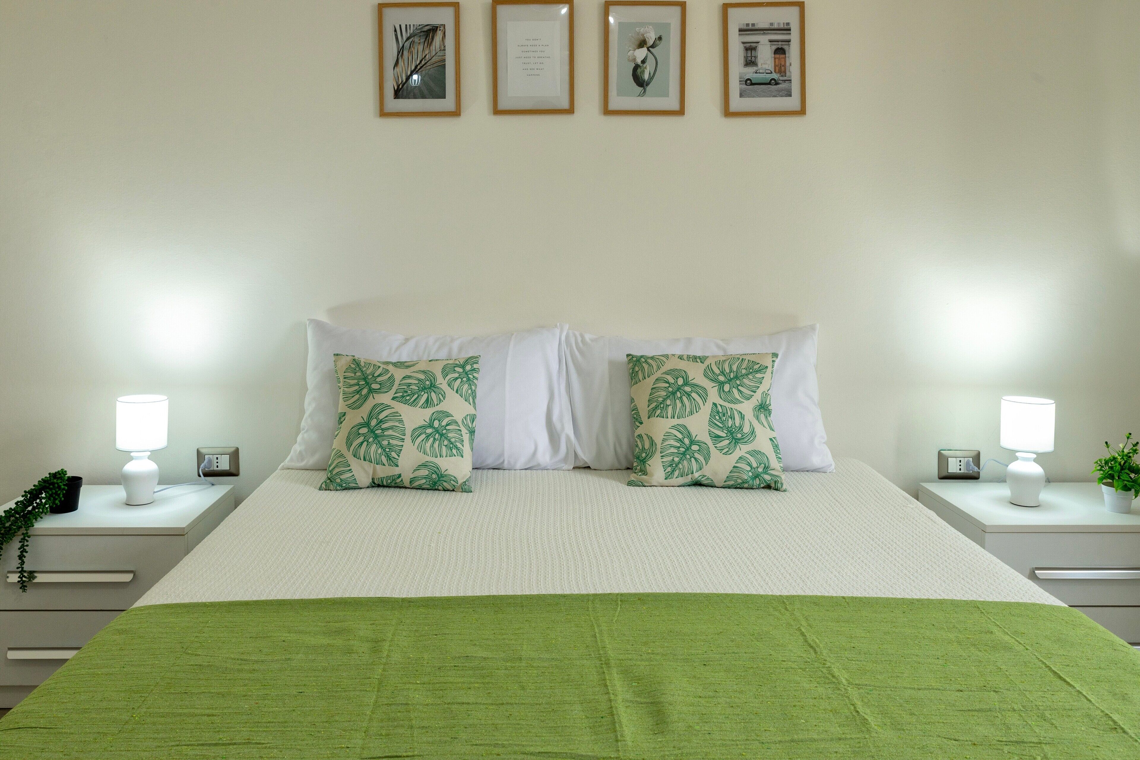 Una cama bien hecha con almohadones blancos y verdes estampados, una manta verde y una mesita de noche con una lámpara y un reloj.