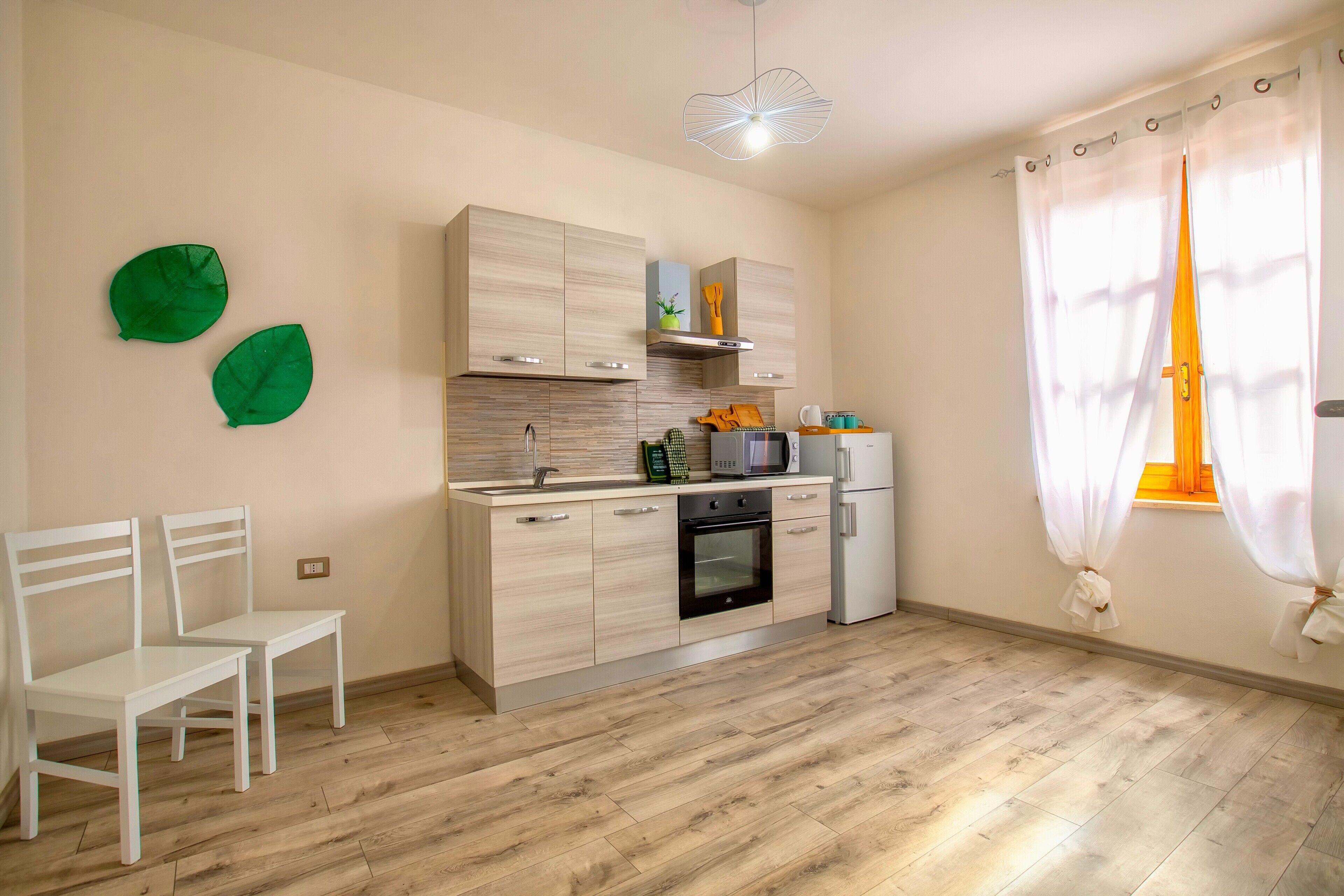 Una cocina con armarios de madera, un microondas y una estufa. Hay dos sillas blancas y una ventana con cortinas.