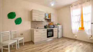 Una cocina con armarios de madera, un microondas y una estufa. Hay dos sillas blancas y una ventana con cortinas.