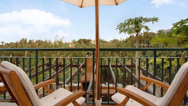 A balcony with two wooden lounge chairs, a large umbrella, and a view of a garden and distant hills.