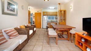 A living room with a sofa, a wooden dining table, a television, and a kitchen in the background.