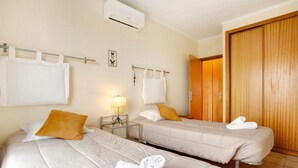 A hotel room with two beds, a wooden wardrobe, a wall-mounted air conditioner, and a small bedside table with a lamp.