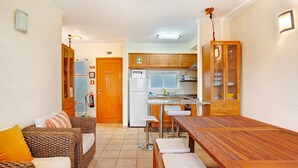 A compact kitchen with a refrigerator, microwave, and wooden cabinets. There is a dining table with chairs and a sofa with cushions.