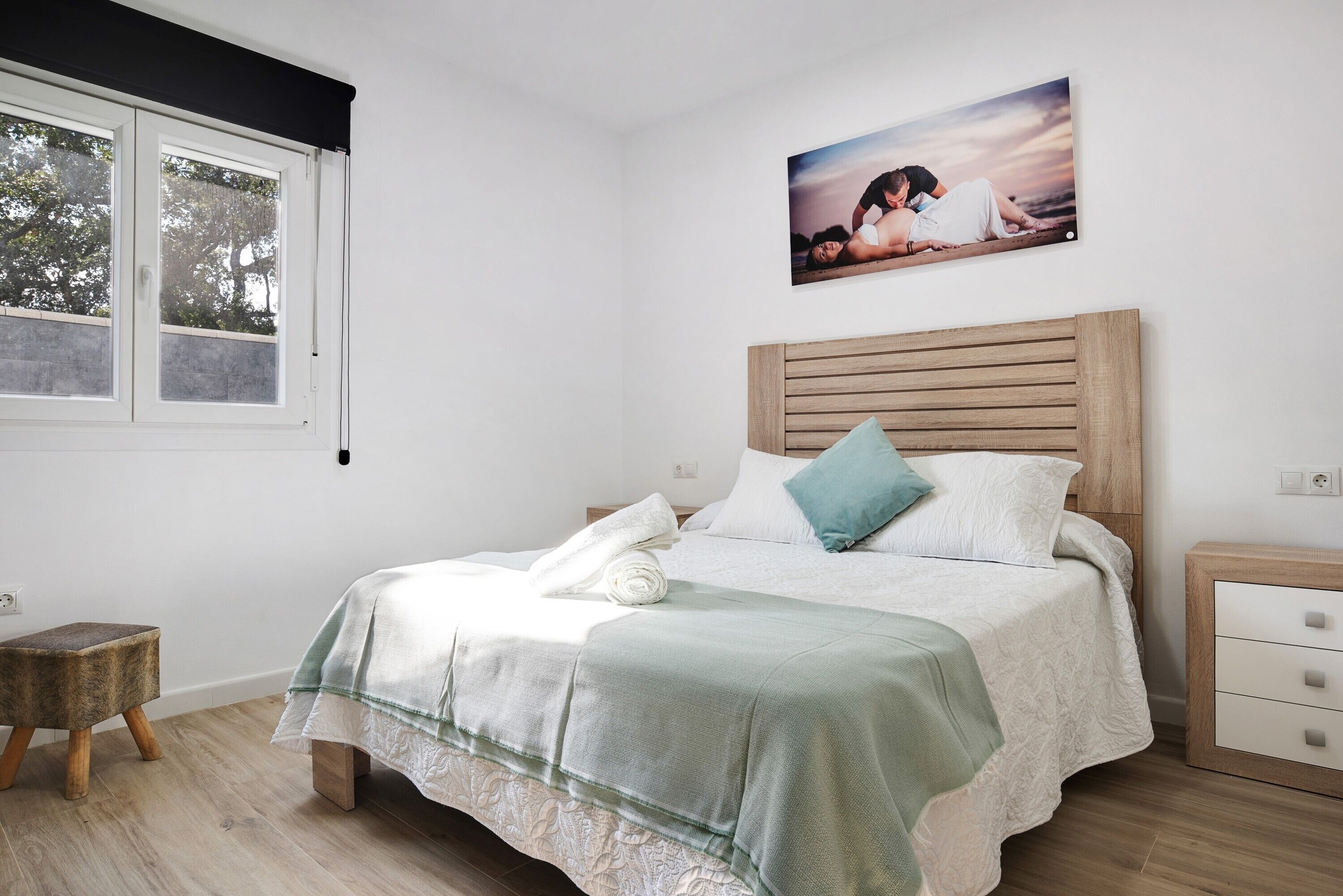 A bedroom with a bed, a nightstand, a small stool, a window with blinds, and a picture on the wall.