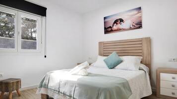 A bedroom with a bed, a nightstand, a small stool, a window with blinds, and a picture on the wall.