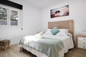 Ein Schlafzimmer mit Bett, Nachttisch, Hocker, Fenster mit Jalousien und einem Bild an der Wand.