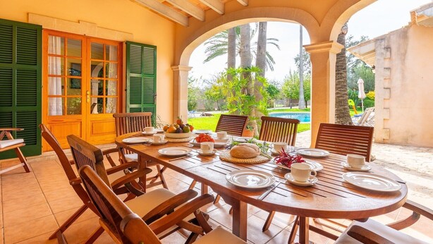 Un área de comedor al aire libre techada con una mesa dispuesta para cuatro personas, rodeada de sillas y vistas a una piscina y palmeras.