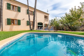 A swimming pool with a tiled edge, a building with green shutters, and a tree in the background.