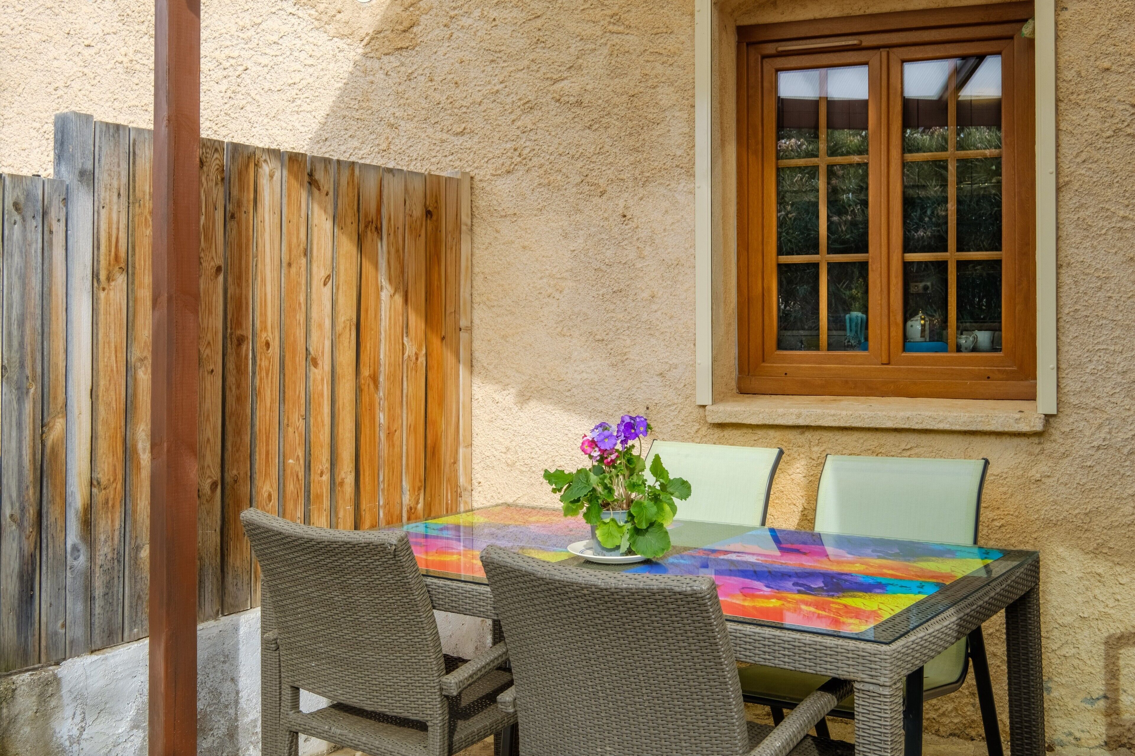 Ein Tisch mit Glasplatte, Korbstühle, ein Holzzaun und ein Fenster mit Blick ins Grüne.