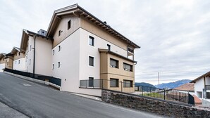 A multi-story building with a flat roof, a stone wall in front, and a paved road in the foreground. - Apartment 'Klara' with Mountain View, Private Terrace and Wi-Fi (Mölten)