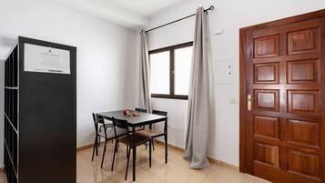 A room with a wooden door, a window with curtains, a table with chairs, and a black cabinet.