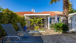 Une terrasse en bois avec des chaises longues, un patio couvert et une maison avec un toit recouvert de tuiles rouges.