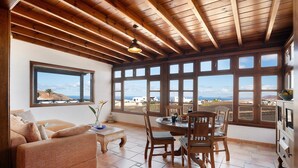 A room with a wooden ceiling, large windows with a view, a sofa, a dining table with chairs, and a small side table. - Vacation Home "Finca Acequia" with Sea View, Private Pool & Wi-Fi (Tinajo)