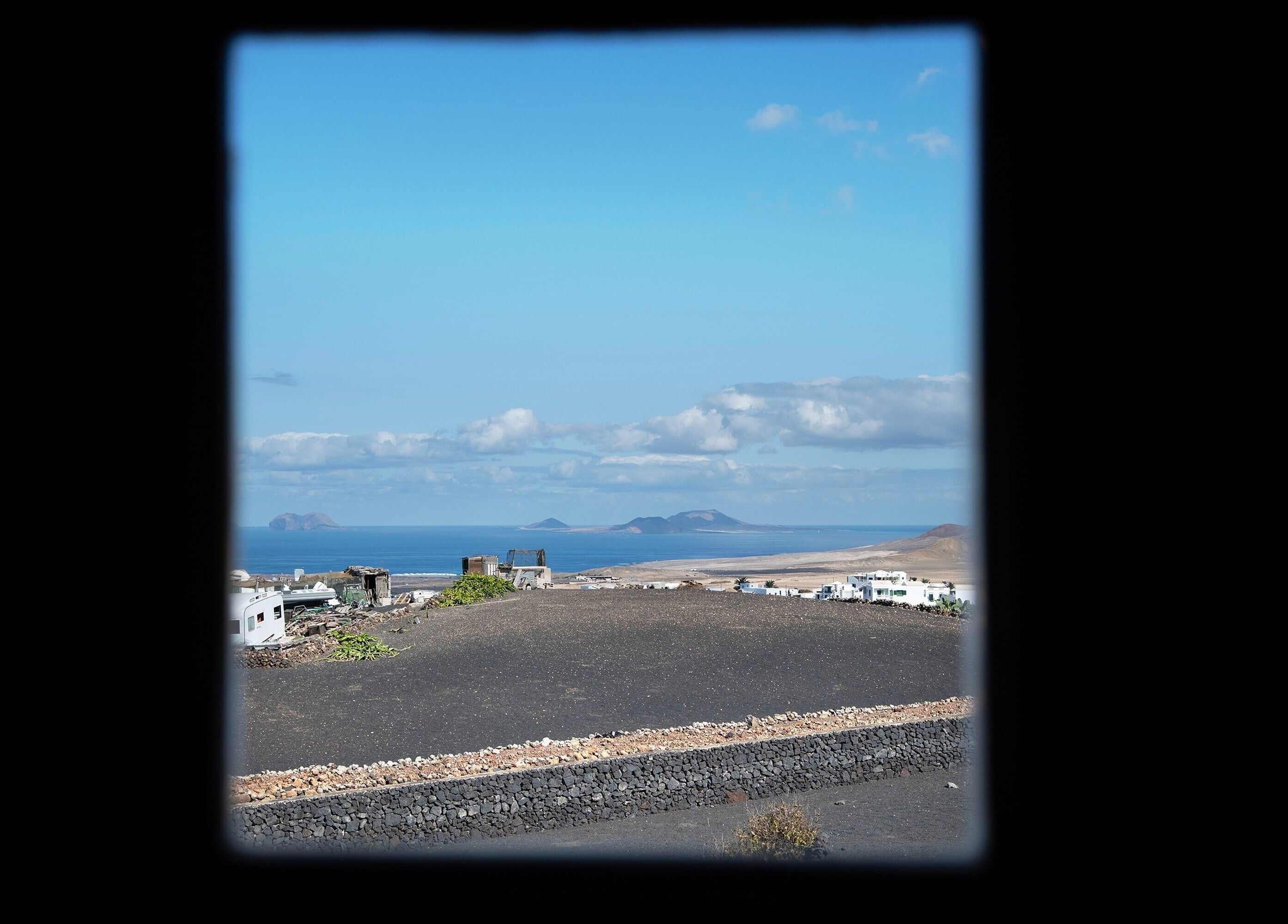 A coastal landscape with a clear sky, distant mountains, and a small settlement with white buildings.