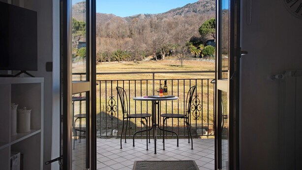 Eine offene Tür zu einem Balkon mit Tisch und Stühlen, Blick auf eine gebirgige Landschaft und eine Uhr an der Wand.
