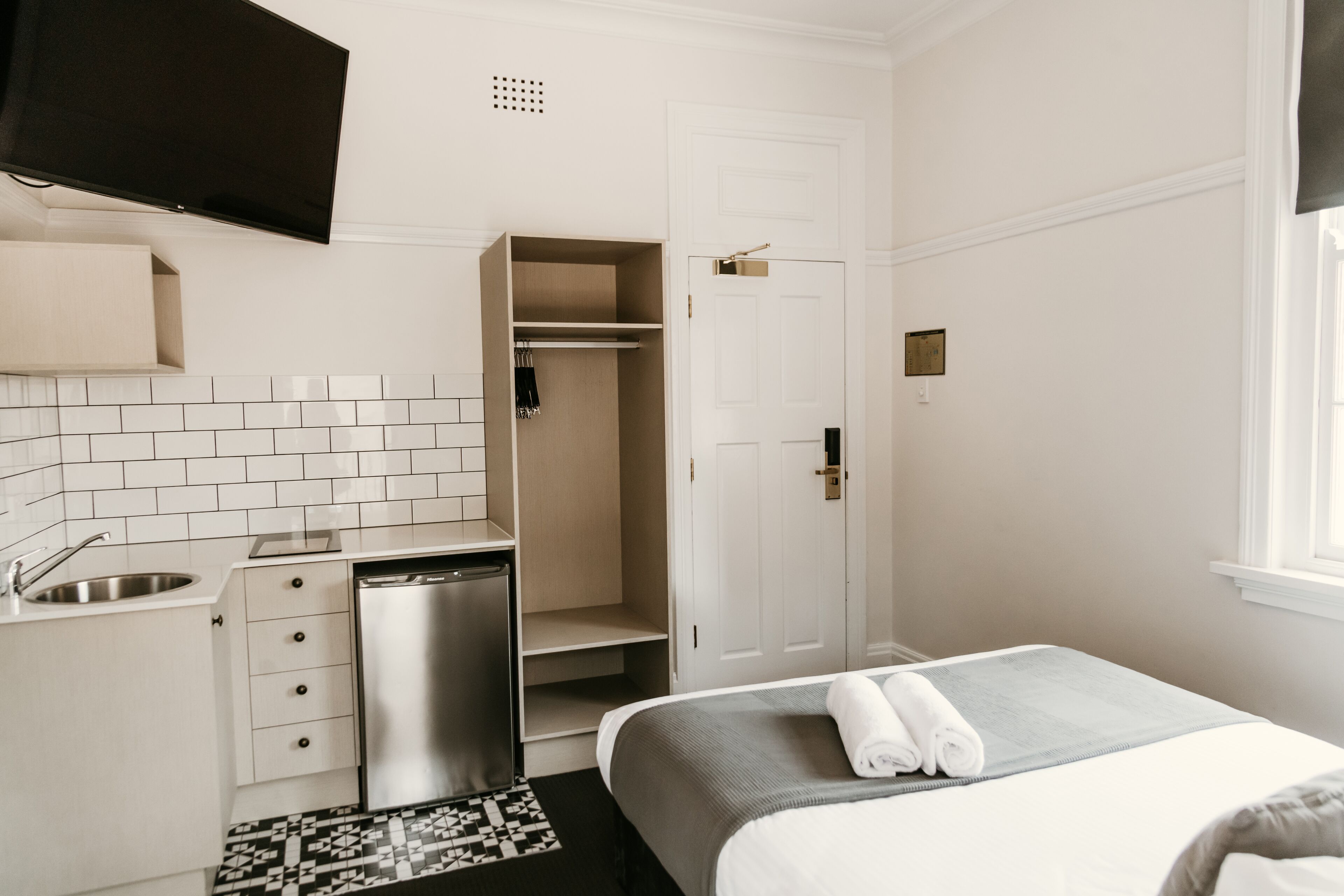 Et hotelværelse med tekøkken, en seng med et pænt foldet håndklæde, en hvid dør og et vindue.