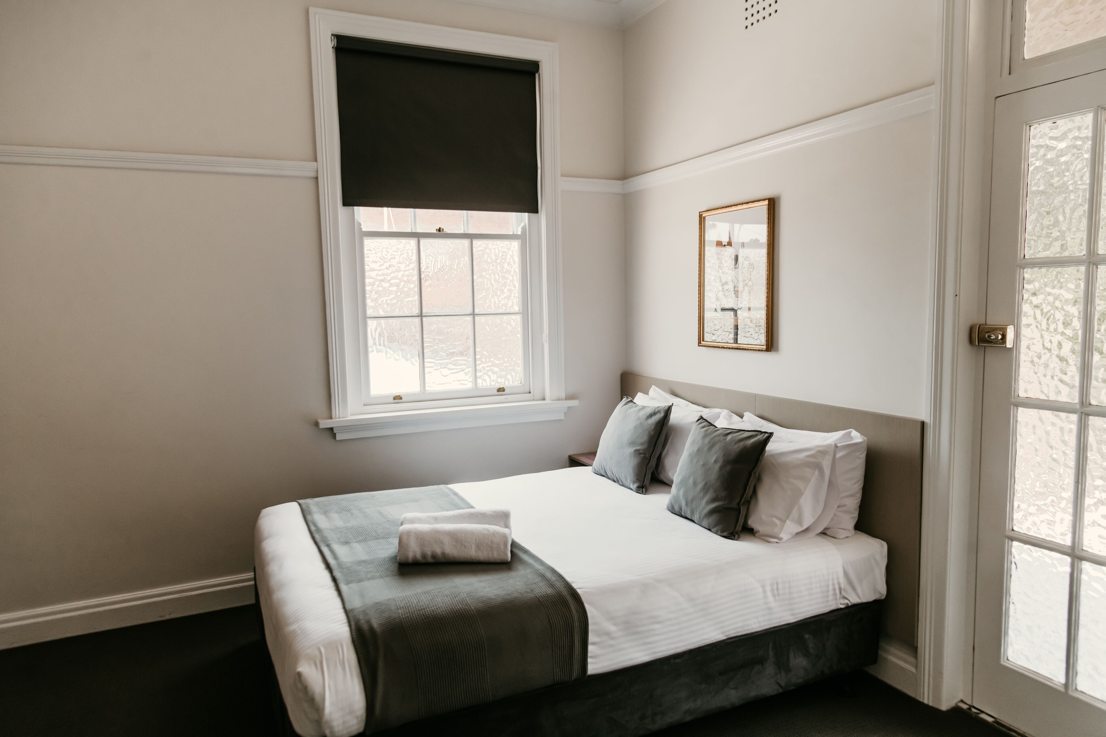 A bedroom with a bed, a window with a dark blind, a framed picture on the wall, and a door with glass panels.