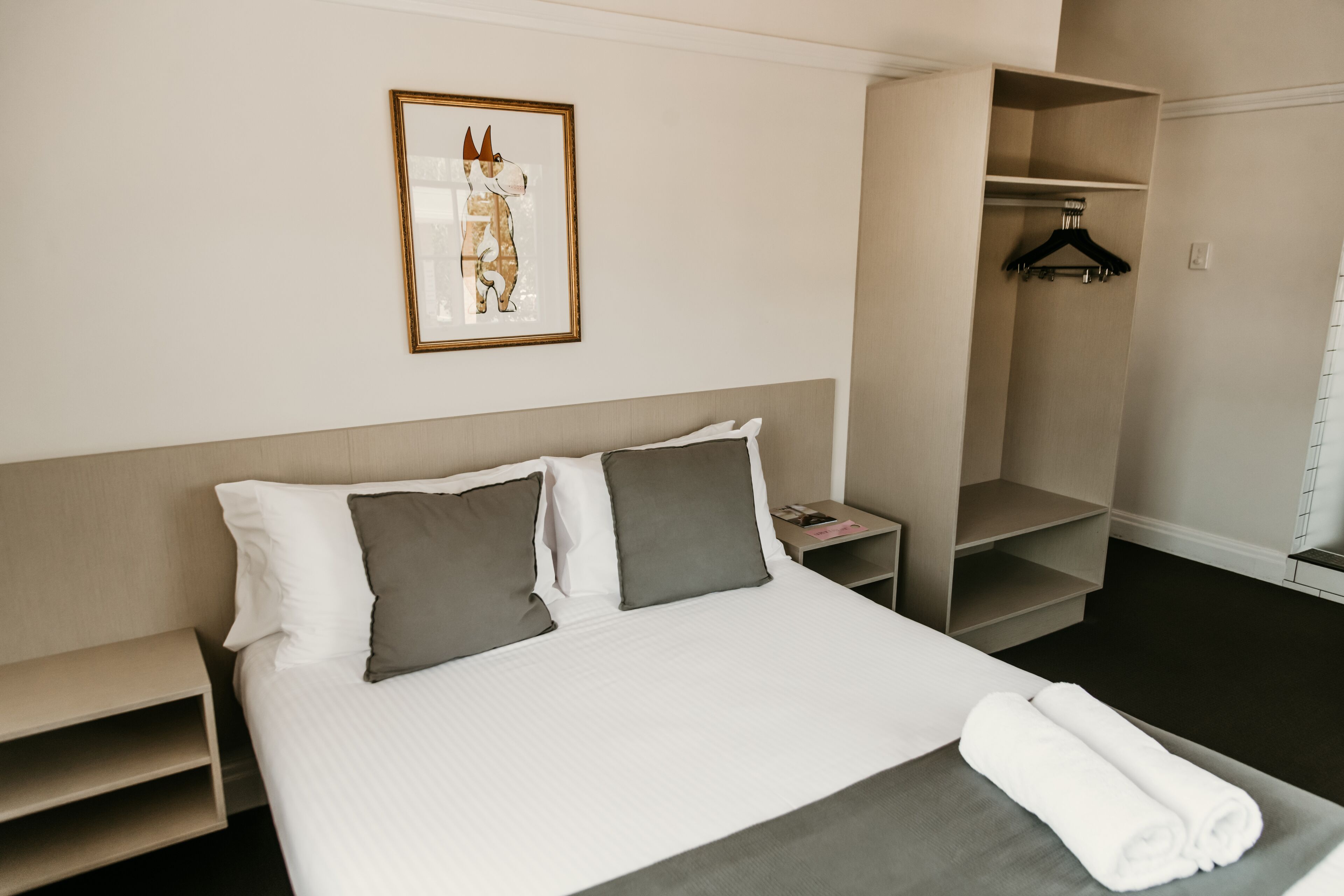 Ein Hotelzimmer mit Bett, Kissen, Nachttisch, einem gerahmten Bild, einem Kleiderschrank und einem Handtuch auf dem Bett.