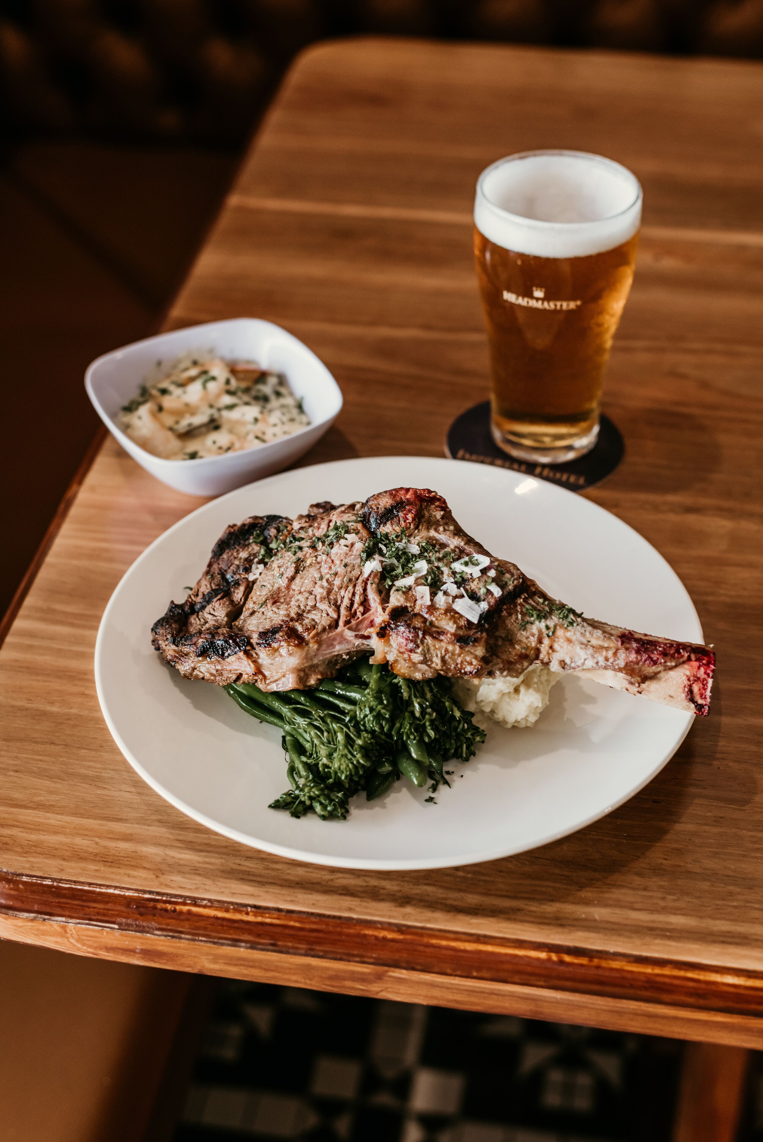 A plate with grilled meat and greens, a glass of beer, and a small bowl of potatoes on a wooden table.