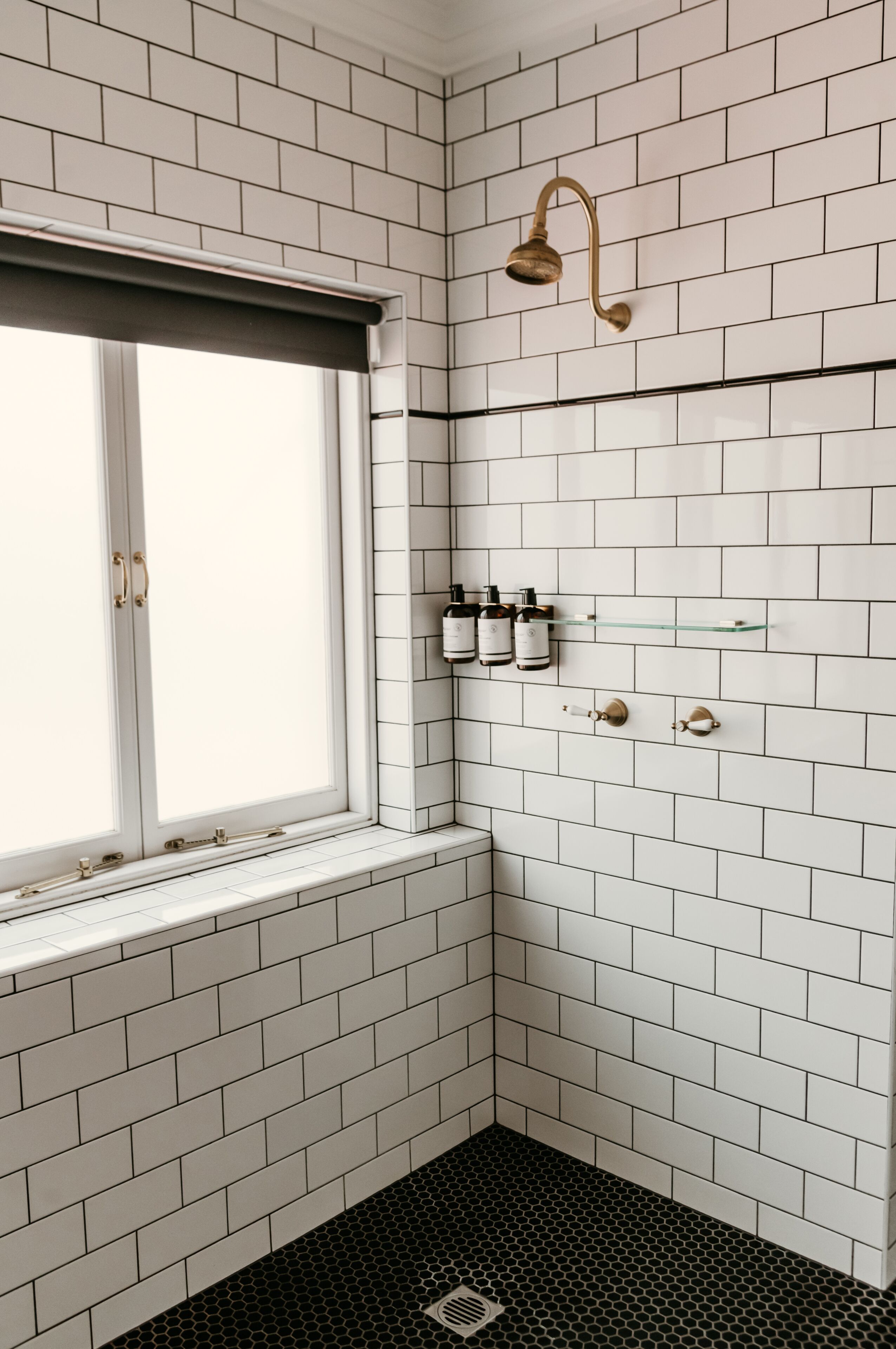 A bathroom with white subway tiles, a shower with a gold showerhead, a window with a white blind, and a shelf with toiletries.