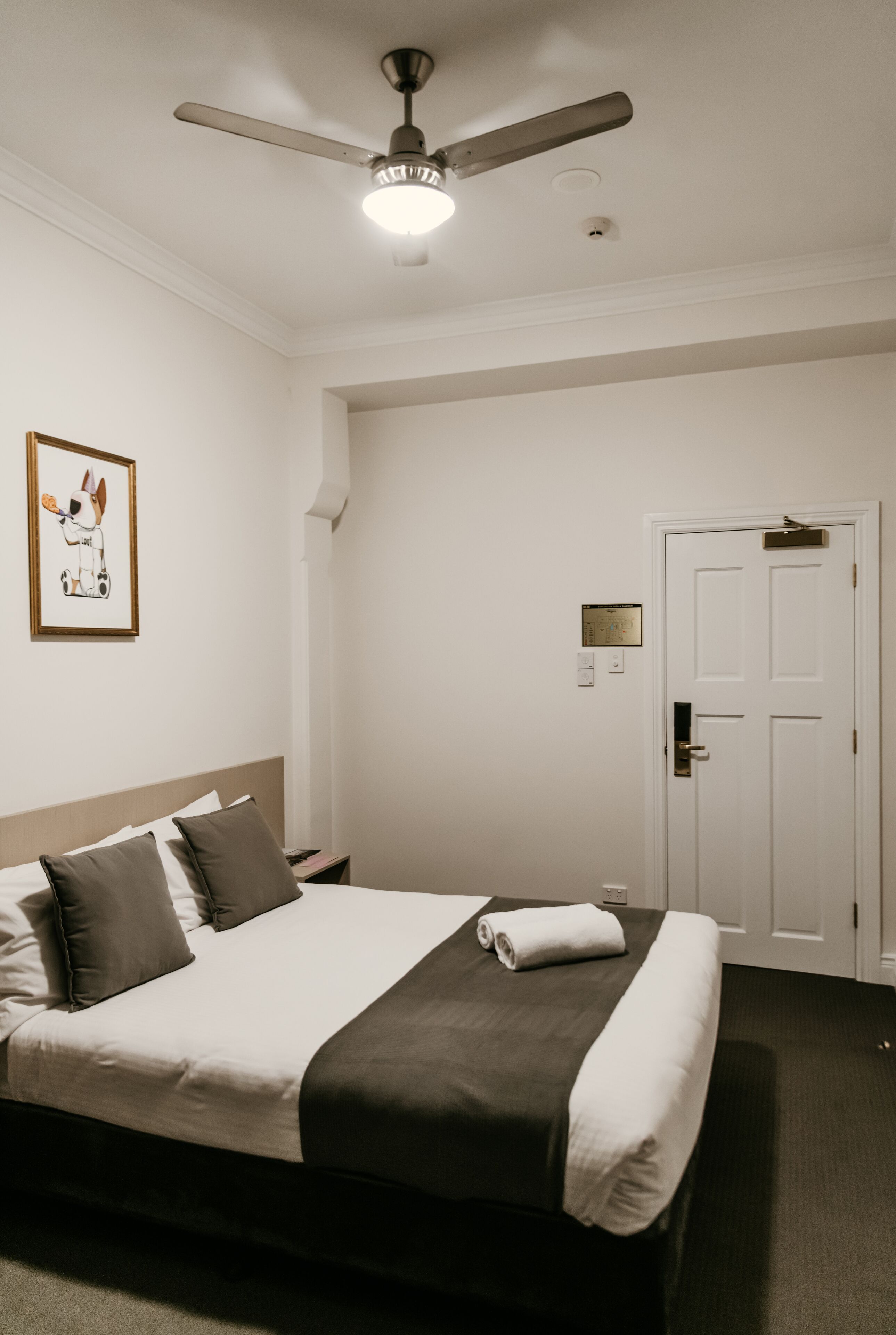 A hotel room with a bed, a ceiling fan, a framed picture on the wall, a door, and a thermostat.
