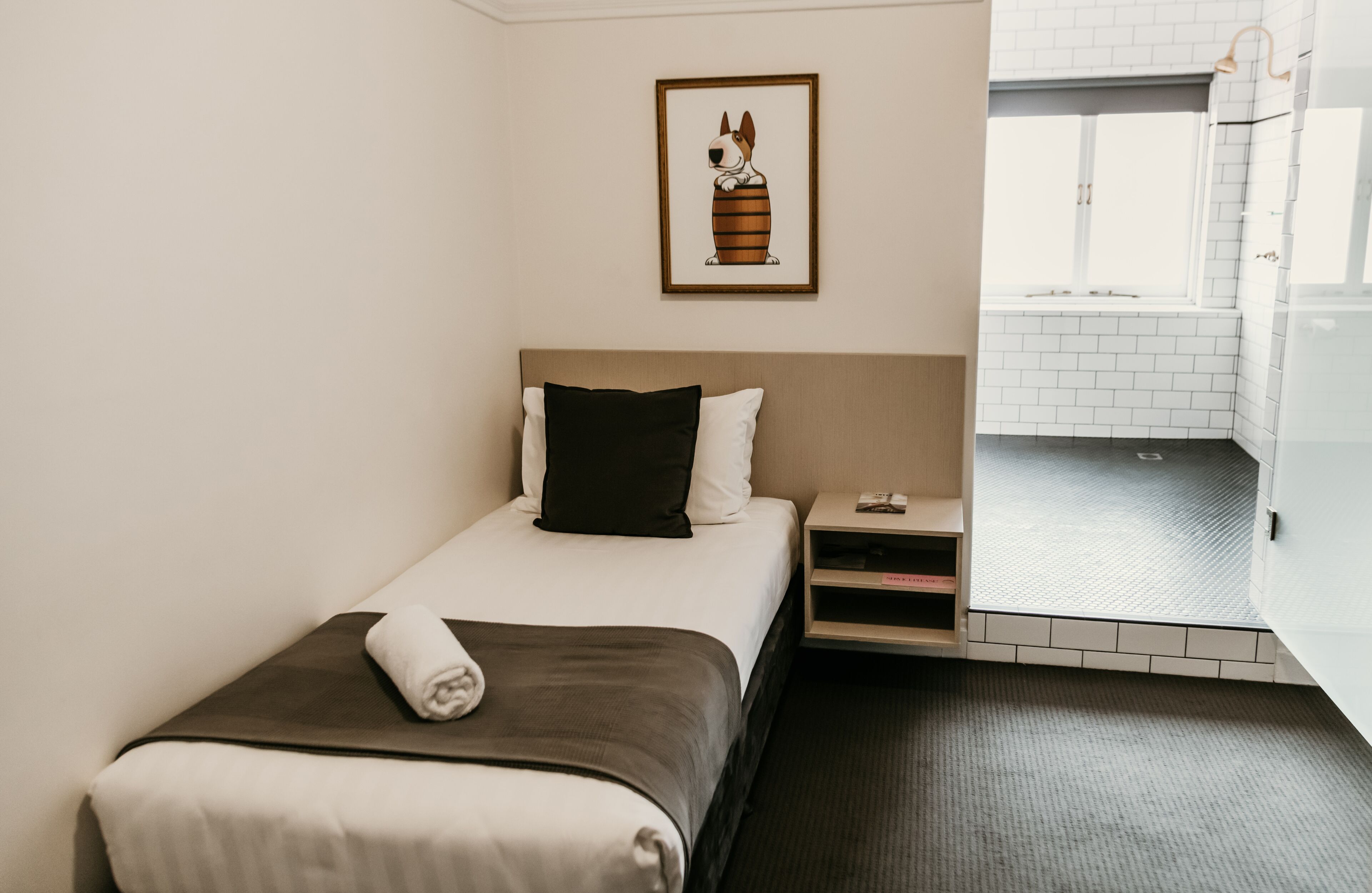 A hotel room with a bed, a nightstand, a framed picture on the wall, and a view of the bathroom.
