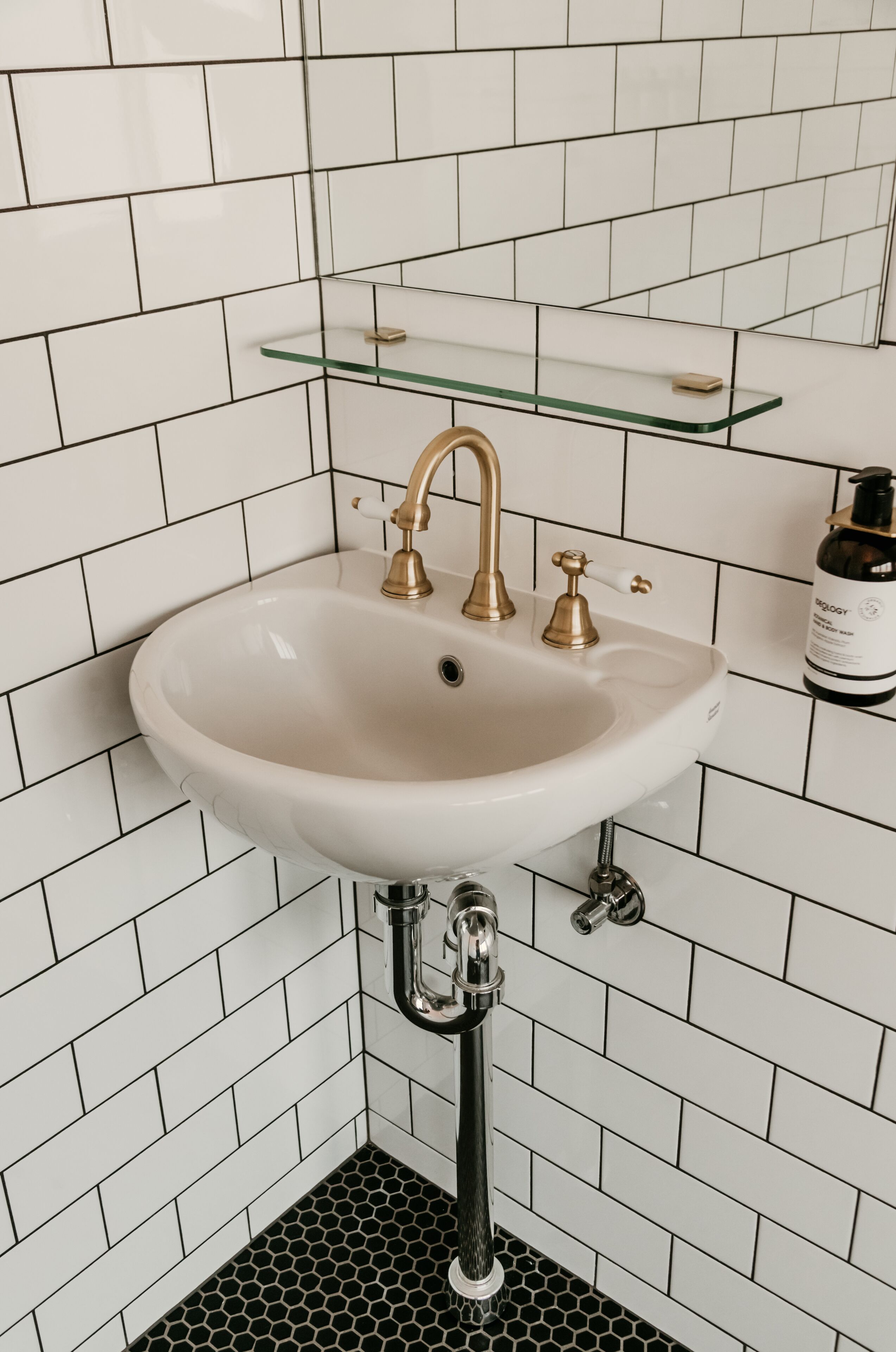 A bathroom with a white sink, gold faucets, and a glass shelf above it.