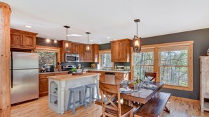 A modern kitchen with wooden cabinets, a stainless steel refrigerator, a central island with stools, and large windows offering a view of the outdoors. - 'Burton on the Rocks' Lake Retreat w/ Hot Tub! (Clayton)