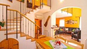 Ein Essbereich mit einem Holztisch, Stühlen und einer grünen Tischdecke. Eine Treppe mit Holzbodenstufen und schwarzen Geländern führt zu einer oberen Ebene.