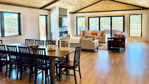 Un'ampia zona giorno con alto soffitto in legno, ampie finestre che offrono vista su un paesaggio montano, un camino e un'area pranzo con mobili in legno.