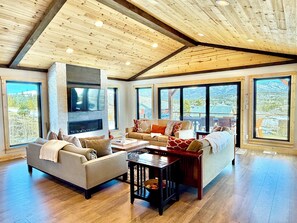 A spacious living room with a fireplace, large windows, and a high ceiling with exposed beams.