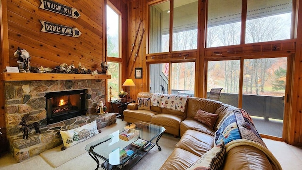 Un acogedor interior de cabaña con chimenea de piedra, sofá de cuero y ventanas amplias con vistas al bosque.
