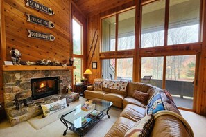 Un acogedor interior de cabaña con chimenea de piedra, sofá de cuero y ventanas amplias con vistas al bosque.