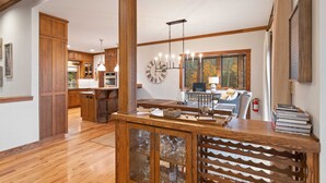 A spacious living area with wooden flooring, a central wooden table, and a large window with a view of trees. - Mountain Home in Private Gated Community (Boone)