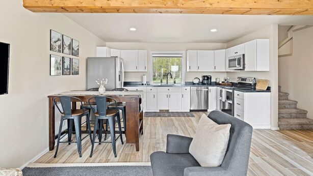 Una cocina moderna con armarios blancos, un comedor con mesa y sillas de madera, y un sofá gris.