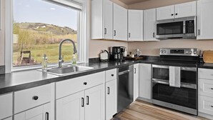 Una cocina moderna con armarios blancos, un lavamanos de acero inoxidable, un microondas, un horno y una ventana con vistas a un paisaje cubierto de césped.